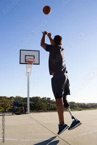 Young man with leg prosthesis throwing basketball into hoop. Back view of male basketball player with disability jumping while playing game outside. Wheelchair in background. Amputee sports concept © KAMPUS
