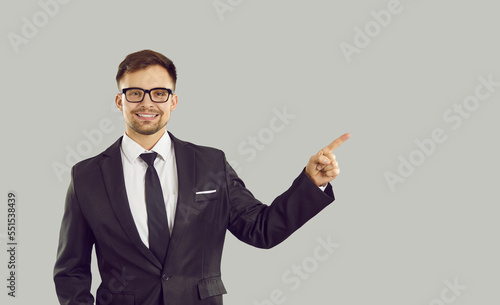 Young successful Caucasian man pointing with finger at copy space next to him with smile recommending business solution or consulting service dressed in jacket and tie stands on studio background