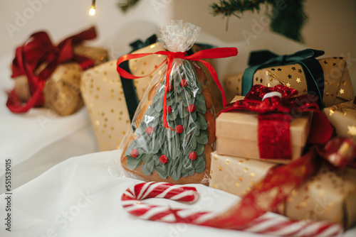 Stylish christmas gifts with cookie and candy cane under christmas tree with golden lights. Wrapped christmas presents in golden paper with red and green ribbons in festive room. Merry Christmas