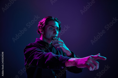 Portrait of young man in casual black shirt nervously looking isolated over gradient dark purple background in neon light. Concept of human emotions, facial expression.