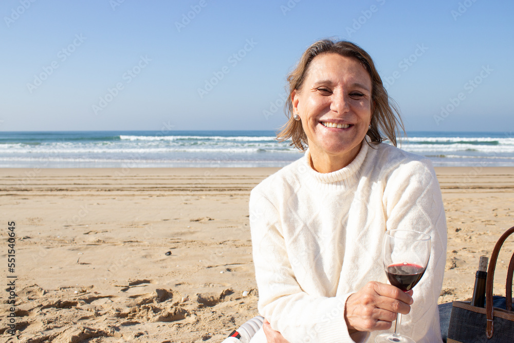 Happy senior woman drinking wine at seashore while having picnic on sunny day. Charming blonde middle-aged lady holding glass of red wine and looking at camera. Retirement, lifestyle, tourism concept