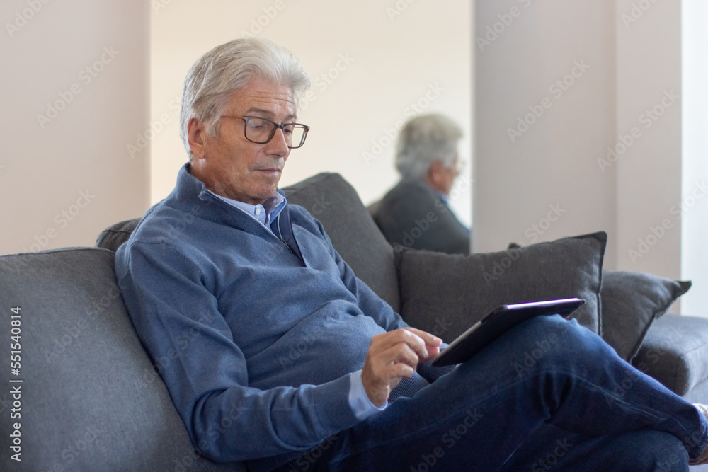Serious elderly businessman working with tablet. Gray-haired man in glasses sitting on sofa and reading news online. Technology concept Serious elderly businessman working with tablet. Gray-haired man in glasses sitting on sofa and reading news online. Technology concept