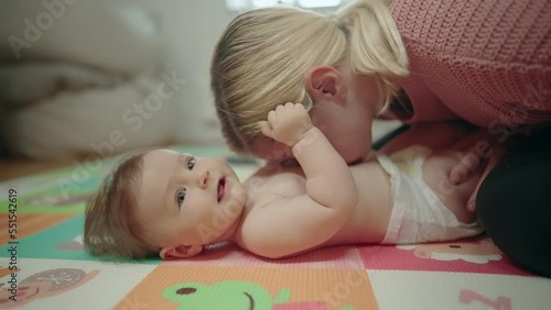 Slow motion - Mother blowing on her daughter's tummy and tickling her while baby holds her hair, both lying on a colorful mat on the floor at daylight photo
