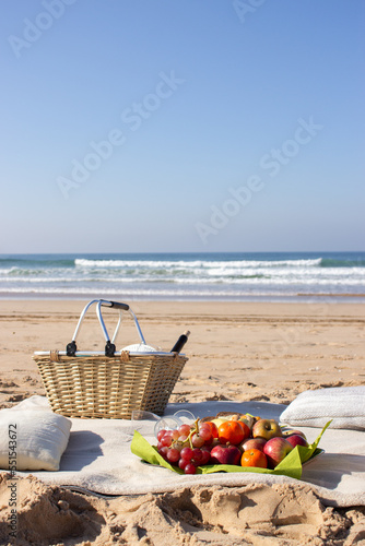 Empty picnic place at sandy seashore on sunny day. Basket, pillows, food and wine glasses on beige blanket. Fantastic seascape background with fleecy waves rushing to shore. Leisure, food concept