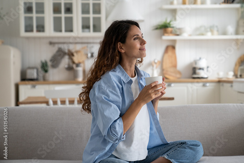 Obraz na plátne Pleased smiling woman resting on sofa with cup of tea enjoying alone time at home, looking aside with dreamy face expression
