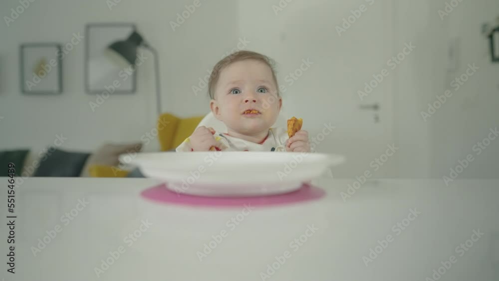 Medium shot - A baby girl sitting in front of a table, wearing a polka-dotted bib, eating food and making funny faces Medium shot - A baby girl sitting in front of a table, wearing a polka-dotted bib, eating food and making funny faces