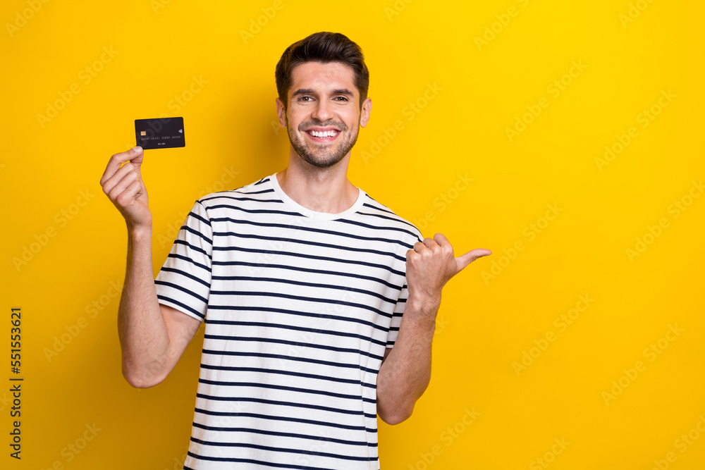 Photo of cheerful nice man toothy smile hold debit card direct finger empty space isolated on yellow color background Photo of cheerful nice man toothy smile hold debit card direct finger empty space isolated on yellow color background