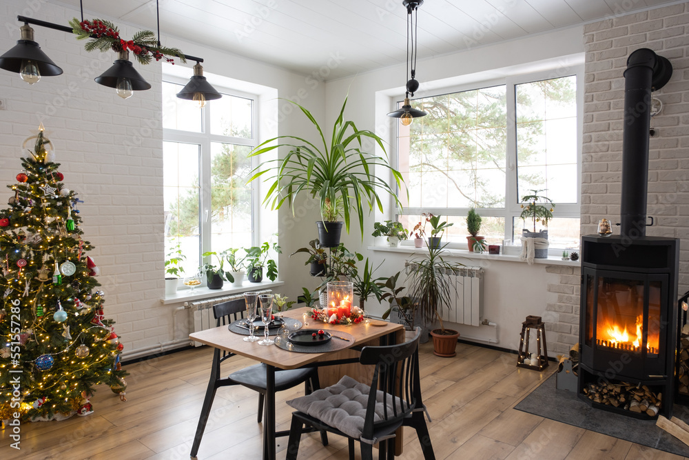 Fototapeta premium Festive interior of house is decorated for Christmas and New Year in loft style with black stove, fireplace, Christmas tree. Warm studio room with set table, burning wood, cozy and heating of home
