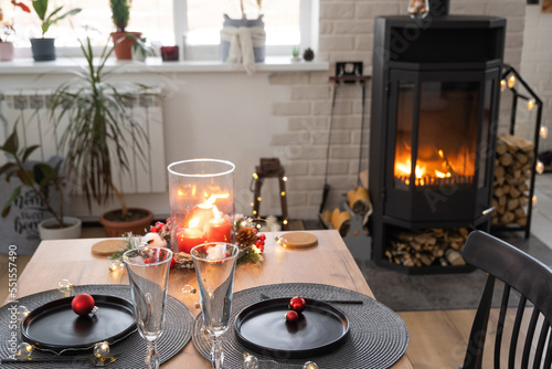Festive interior of house is decorated for Christmas and New Year in loft style with black stove, fireplace, Christmas tree. Warm studio room with set table, burning wood, cozy and heating of home