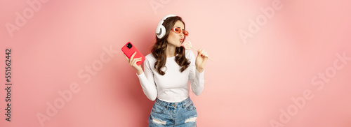 Summer girl laughing, eathing lolipop and listening music in headphones, dancing with smartphone against pink background photo