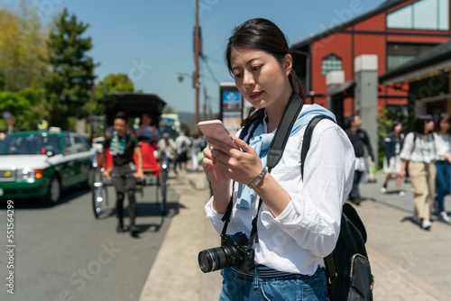 concentrated asian Japanese woman traveler looking at online travel guide on mobile phone at the roadside of Arashiyama Mountains street in Kyoto japan with a man pulling rickshaw at background