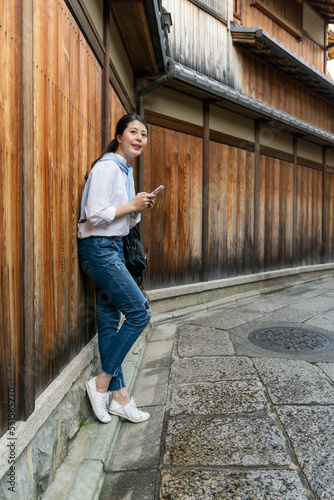 full length of cheerful asian Japanese woman visitor learning about Gion Ishibeikoji lane on phone while traveling to Kyoto japan. she leans against wooden wall and admiring the historic alley © PR Image Factory