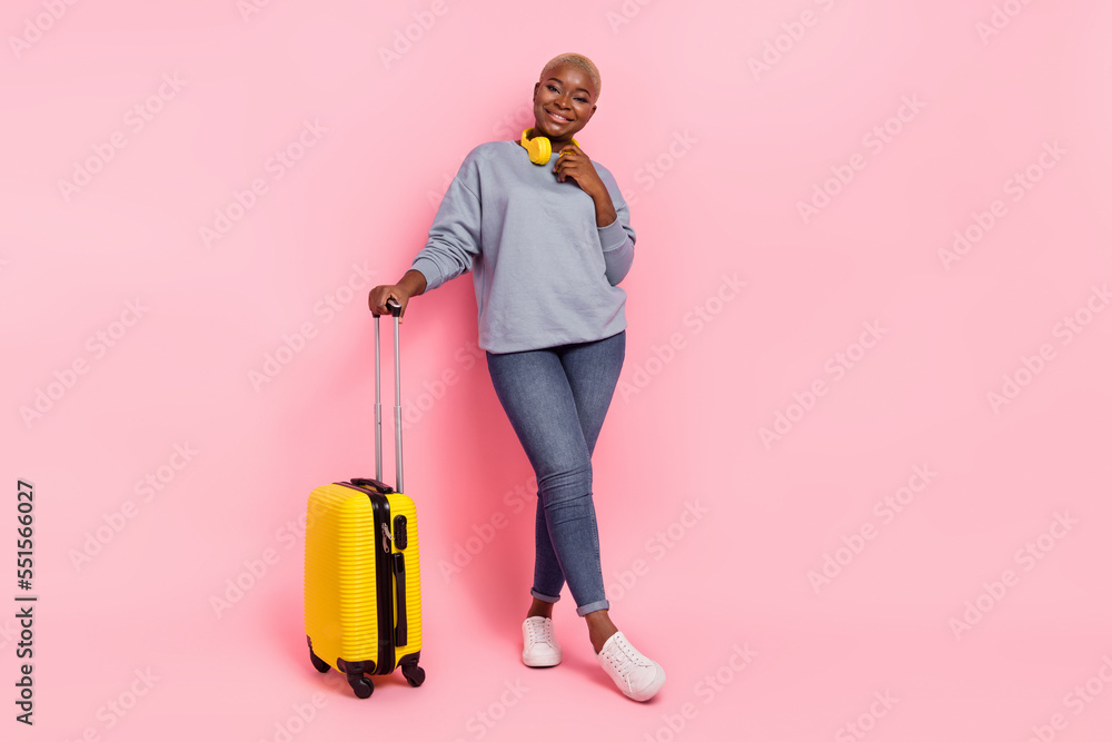 Full size portrait of pretty positive lady hand touch wireless headphones hold suitcase isolated on pink color background Full size portrait of pretty positive lady hand touch wireless headphones hold suitcase isolated on pink color background