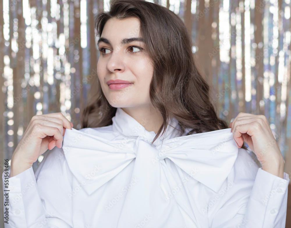 Young happy woman holding festive bow on her white blouse on the tinsel foil background. Christmas or New Year celebration party fashion concept portrait Young happy woman holding festive bow on her white blouse on the tinsel foil background. Christmas or New Year celebration party fashion concept portrait