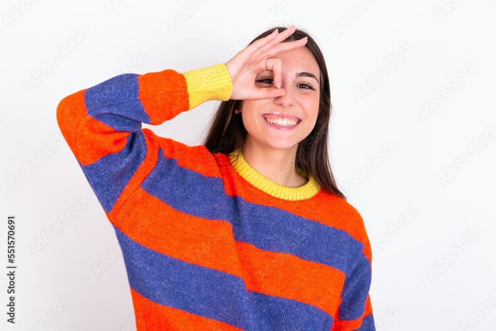 Young caucasian woman wearing colorful knitted sweater over white background doing ok gesture with hand smiling, eye looking through fingers with happy face. Young caucasian woman wearing colorful knitted sweater over white background doing ok gesture with hand smiling, eye looking through fingers with happy face.