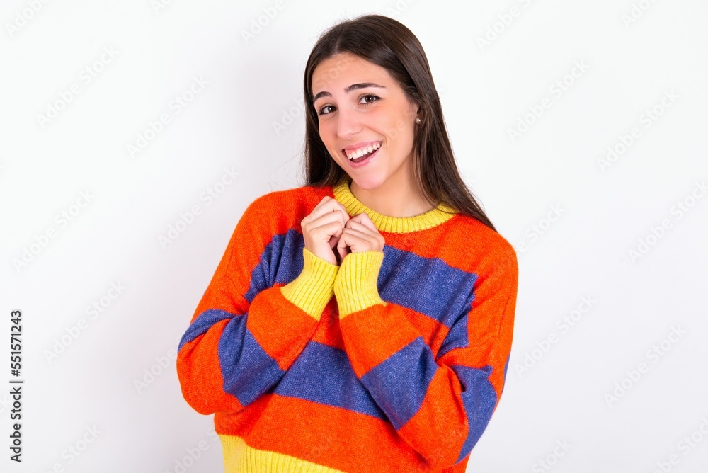 Positive Young caucasian woman wearing colorful knitted sweater over white background smiles happily, glad to receive pleasant news from interlocutor, keeps hands together. People emotions concept. Positive Young caucasian woman wearing colorful knitted sweater over white background smiles happily, glad to receive pleasant news from interlocutor, keeps hands together. People emotions concept.