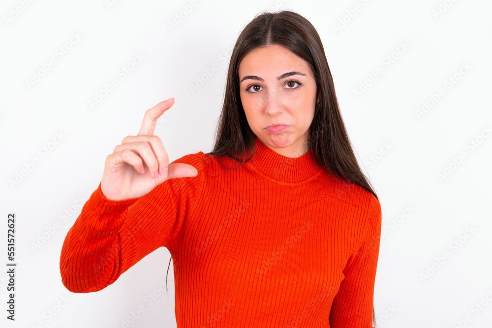 Young caucasian woman wearing red sweater over white background purses lip and gestures with hand, shows something very little. Young caucasian woman wearing red sweater over white background purses lip and gestures with hand, shows something very little.