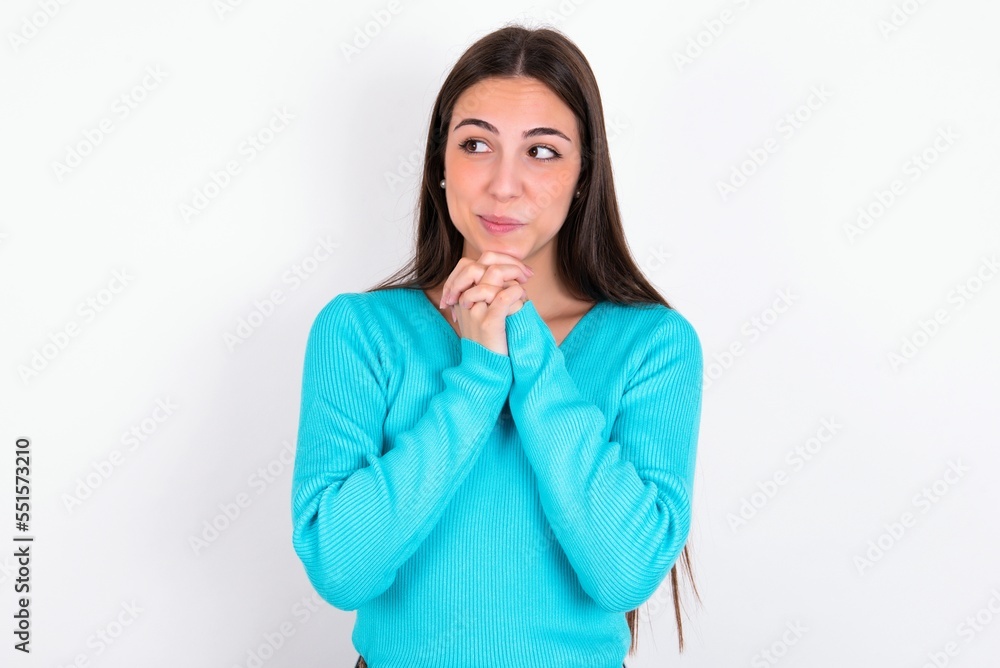 Curious Young caucasian woman wearing blue sweater over white background keeps hands under chin bites lips and looks with interest aside. Curious Young caucasian woman wearing blue sweater over white background keeps hands under chin bites lips and looks with interest aside.