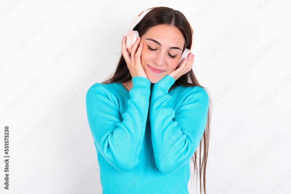 Pleased Young caucasian woman wearing blue sweater over white background enjoys listening pleasant melody keeps hands on stereo headphones closes eyes. Spending free time with music Pleased Young caucasian woman wearing blue sweater over white background enjoys listening pleasant melody keeps hands on stereo headphones closes eyes. Spending free time with music