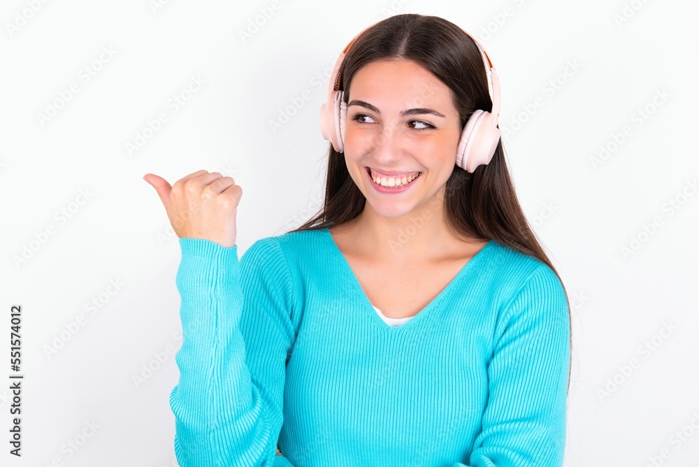 Young caucasian woman wearing blue sweater over white background listens audio track via wireless headphones points thumb away advertises copy space Young caucasian woman wearing blue sweater over white background listens audio track via wireless headphones points thumb away advertises copy space