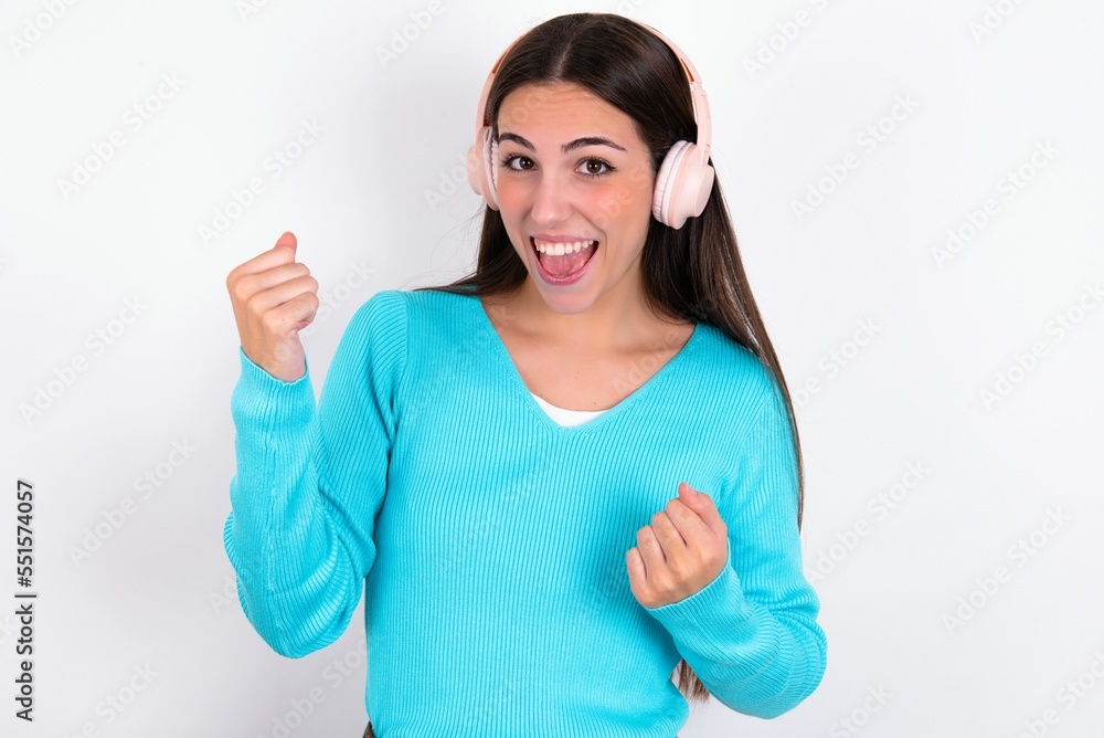 Emotional Young caucasian woman wearing blue sweater over white background exclaims loudly feels like winner raises clenched fists keeps mouth opened wears stereo headphones on ears makes yes gesture Emotional Young caucasian woman wearing blue sweater over white background exclaims loudly feels like winner raises clenched fists keeps mouth opened wears stereo headphones on ears makes yes gesture