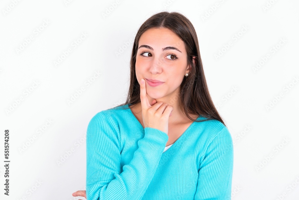 Lovely dreamy Young caucasian woman wearing blue sweater over white background keeps finger near lips looks aside copy space. Lovely dreamy Young caucasian woman wearing blue sweater over white background keeps finger near lips looks aside copy space.