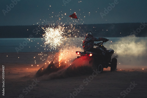Athlete on a quad bike rides at night on a sandy beach with fireworks