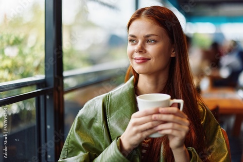Portrait of a woman influencer drinking a drink from a mug in a cafe and smiling with her teeth looking out the window, content blogger close-up