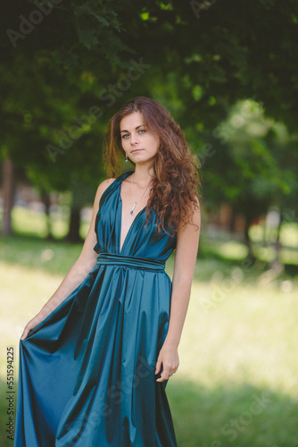 curly haired young woman in fashionable long dress smiling on sunny graduation day, bridesmaid outdoors 