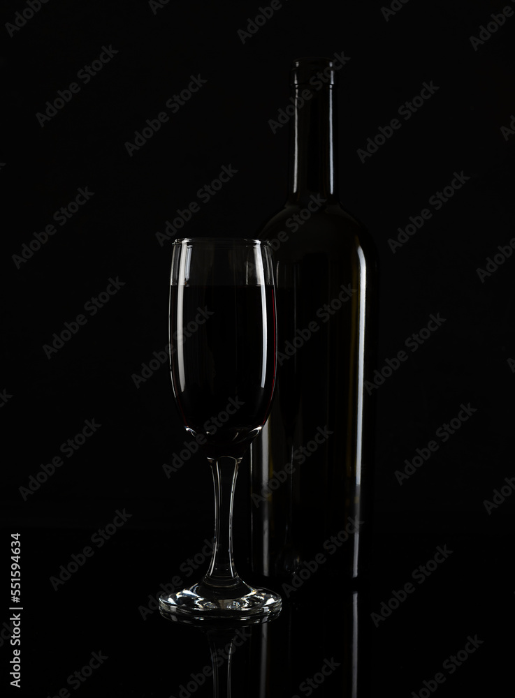 Fototapeta premium A tall glass of red wine and a bottle on a black table. White highlights on the glass and bottle. Black background
