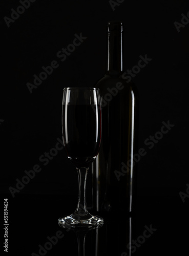 A tall glass of red wine and a bottle on a black table. White highlights on the glass and bottle. Black background