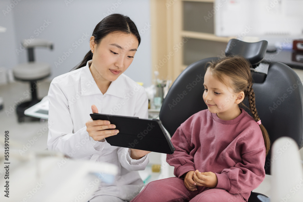 Medicine, pediatric dentistry and oral care concept. Female smiling asian dentist showing tablet pc computer to happy kid patient, caucasian preschool girl at modern dental clinic. Medicine, pediatric dentistry and oral care concept. Female smiling asian dentist showing tablet pc computer to happy kid patient, caucasian preschool girl at modern dental clinic.