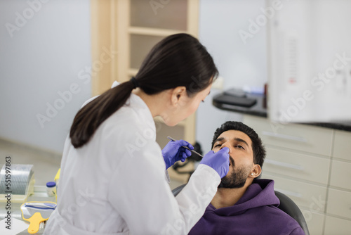 Beautiful smile with white teeth. Asian dentist examines the oral cavity of young bearded handsome man through a magnifying glass in the dental office clinic.
