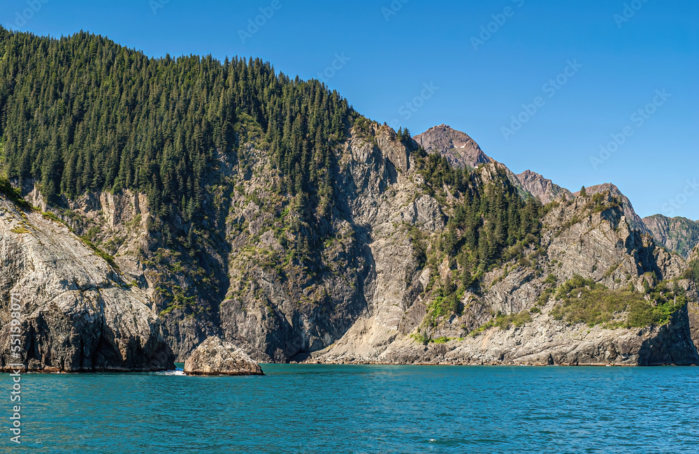 Resurrection Bay, Alaska, USA - July 22, 2011: Sunlighted gray rocky cliffs, green forested on top, descends into azure water under blue sky