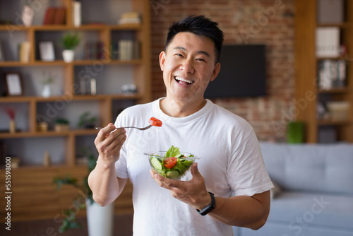 Happy asian mature man holding fork and bowl with fresh vegetable salad, eating healthy lunch after domestic training