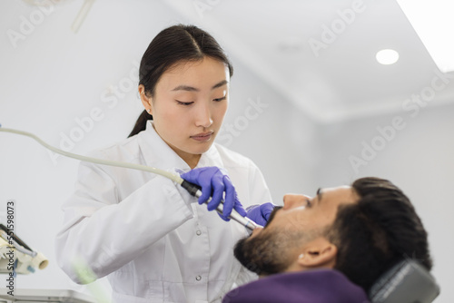 Shot of young pretty female asian dentist in white uniform  making dental check up and caries treatment with drill for her attractive male patient  bearded man  sitting in dental chair at clinic.