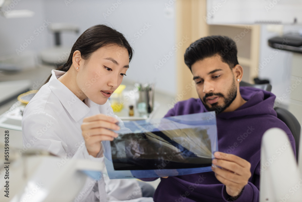Focus on x ray picture, attractive young bearded man, sitting on dentist chair and looking at x-ray scan image of teeth together with his cheerful female asian dentist at clinic. Focus on x ray picture, attractive young bearded man, sitting on dentist chair and looking at x-ray scan image of teeth together with his cheerful female asian dentist at clinic.