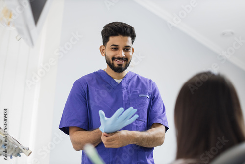Confident bearded male dentist puts on blue rubber gloves, standing in a dental office, ready for the examination and treatment of his female patient. Happy patient and dentist concept