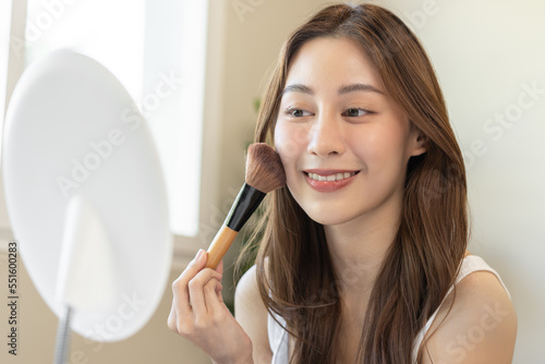 Happy routine beauty concept, pretty asian young woman, girl make up face by applying powder foundation by brush around face, looking at the mirror at home. Female look with natural fashion style.