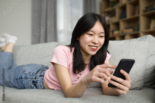 Asian lady enjoy spare time alone, lying on sofa and watching news on cellphone, relaxing at home