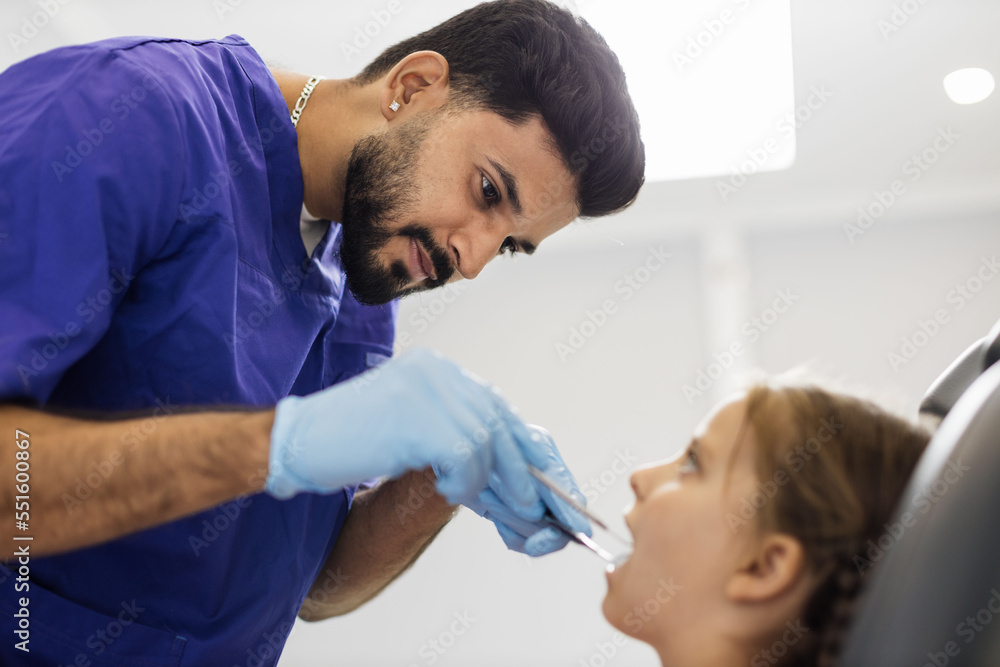 Side low angle view of smiling male professional doctor in blue uniform and gloves, making examination or curing caries and toothache for his little patient, cute kid girl, sitting in dental chair. Side low angle view of smiling male professional doctor in blue uniform and gloves, making examination or curing caries and toothache for his little patient, cute kid girl, sitting in dental chair.