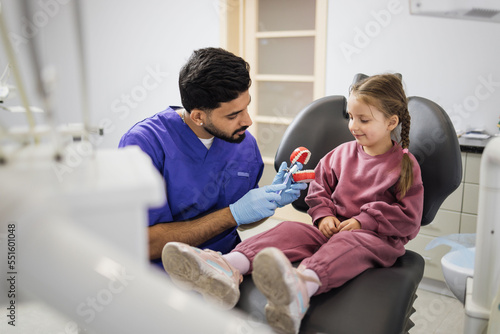 Side view of young confident bearded pediatric dentist showing to little preschool girl how to brush teeth on artificial jaw at light modern dental office. Pediatric dentistry. © sofiko14