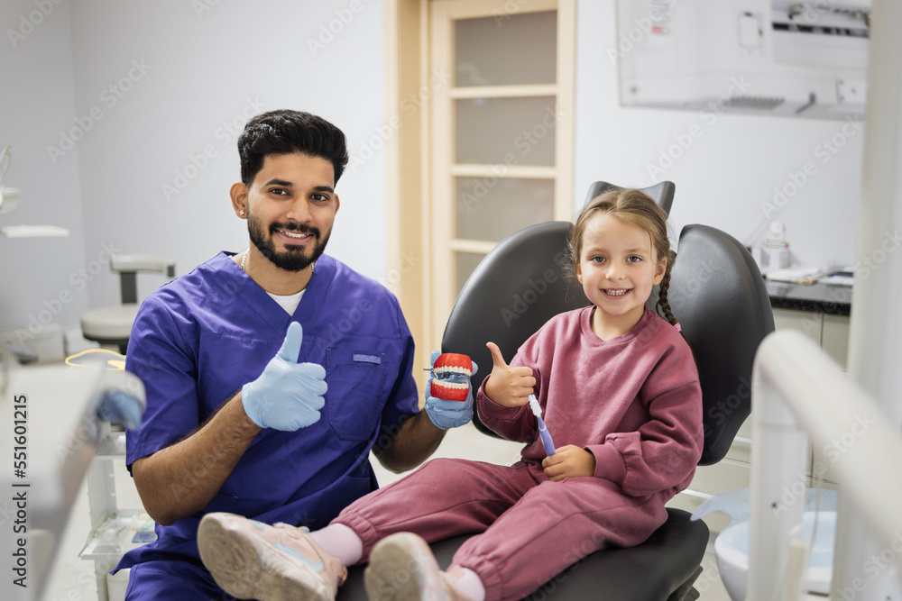 Happy confident male dentist tells little child girl how to brush the teeth on artificial jaw model showing thumbs up. Caries prevention, pediatric dentistry, milk teeth hygiene concept. Happy confident male dentist tells little child girl how to brush the teeth on artificial jaw model showing thumbs up. Caries prevention, pediatric dentistry, milk teeth hygiene concept.