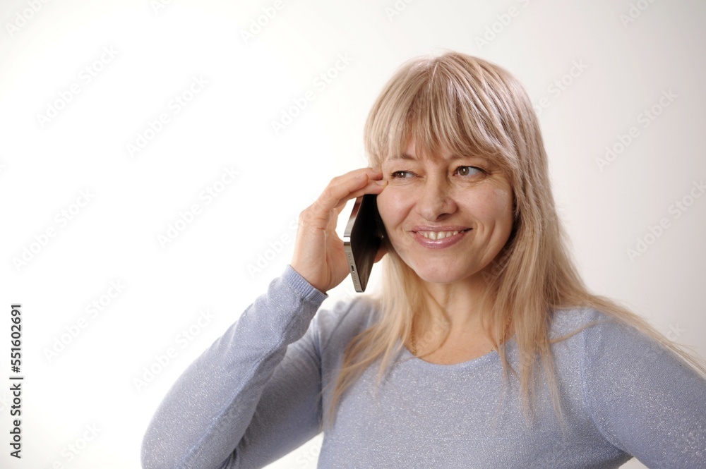 Fototapeta premium An adult woman 45-50 with blond hair in a blue dress is standing with a mobile phone Green screen chroma key She smiles advertising any products supply demand