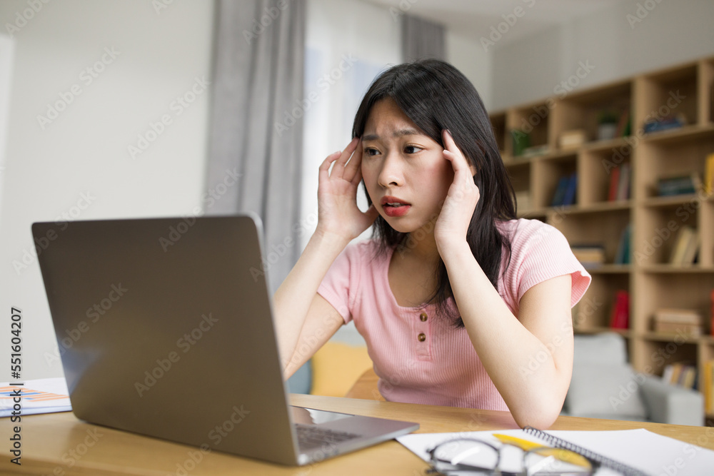 Sad tired asian lady with laptop suffering from headache, pressing hands to temples, sitting at desk in living room Sad tired asian lady with laptop suffering from headache, pressing hands to temples, sitting at desk in living room