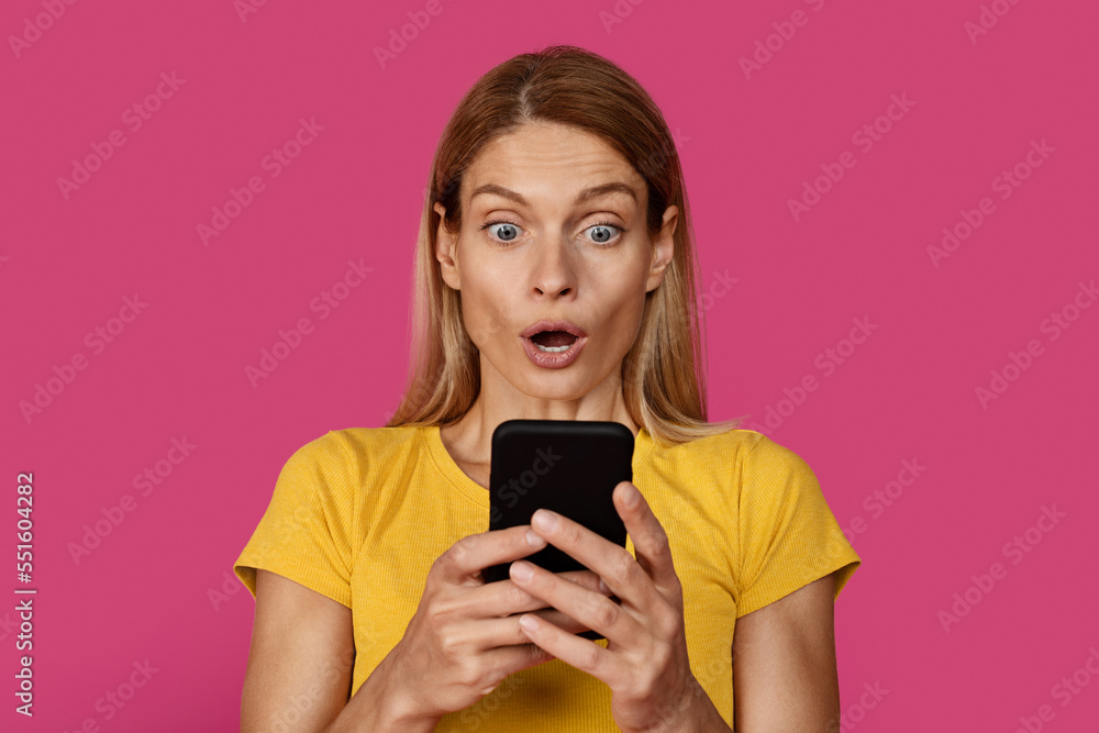 Fototapeta premium Glad surprised mature caucasian blonde female in yellow t-shirt with open mouth looks at smartphone