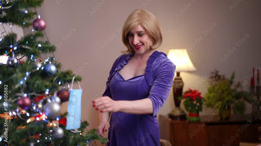 Positive non-binary LGBT woman hanging coronavirus face mask on Christmas tree smiling looking at camera. Portrait of cheerful Caucasian gender-fluid person decorating home on New Year's eve posing Positive non-binary LGBT woman hanging coronavirus face mask on Christmas tree smiling looking at camera. Portrait of cheerful Caucasian gender-fluid person decorating home on New Year's eve posing
