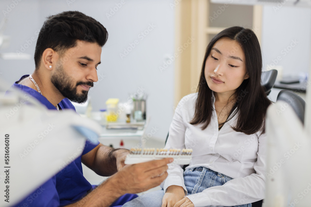 Fototapeta premium Tooth teeth tone palette. Confident bearded male cosmetologist orthodontist dentist choosing selecting color of patient attractive asian woman teeth for whitening at clinic.