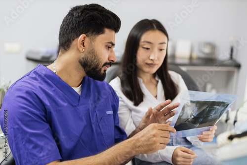 Attractive young asian woman, sitting on dentist chair and looking at x-ray picture of scan image of teeth together with her confident professional male dentist at clinic.