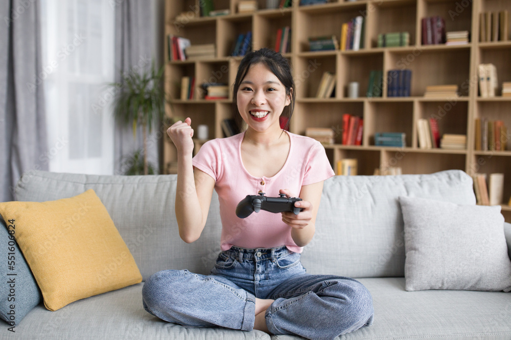 Joyful korean lady with joystick playing online game, raising hand up and celebrating victory in living room interior Joyful korean lady with joystick playing online game, raising hand up and celebrating victory in living room interior